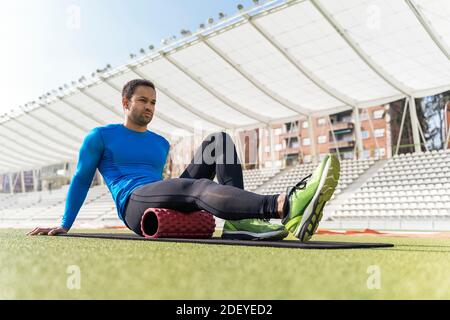 Foto d'inventario di atleta africano-americano seduto sulla pista che allunga le gambe con rullo in schiuma. Foto Stock