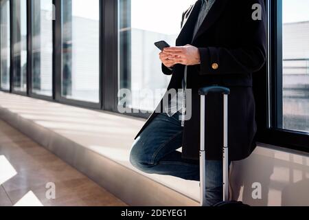 Foto in stock di un closeup di un uomo d'affari irriconoscibile utilizzando il suo telefono in aeroporto. Sta appoggiandosi su una finestra. È serio. Indossa cas Foto Stock