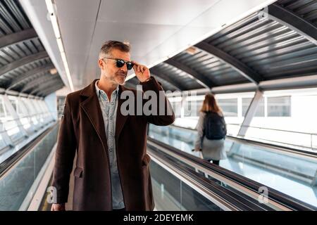 Stock foto di un uomo d'affari caucasico che cammina su un trasportatore a nastro con una valigia che indossa i suoi occhiali da sole per prendere un volo. Sta sorridendo e. Foto Stock