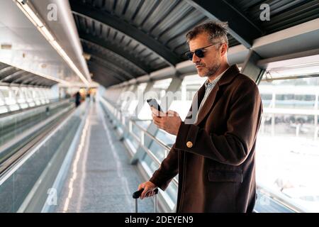 Foto d'inventario di un uomo d'affari caucasico che controlla il suo cellulare prima di prendere un volo in aeroporto. Indossa abiti e occhiali da sole casual. Foto Stock
