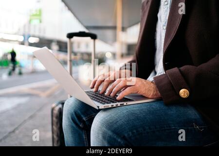 Foto in stock di una vista laterale da vicino di un uomo d'affari irriconoscibile che scrive nel suo laptop. È seduto su una panchina. Ha una valigia accanto a lui. Foto Stock