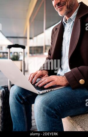 Foto di un primo piano di un uomo d'affari irriconoscibile che scrive nel suo laptop. È seduto su una panchina. Ha una valigia accanto a lui. Foto Stock