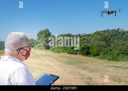 Uomo con capelli grigi che vola un drone su un campo con una tavoletta e una maschera di sicurezza. Nuovo concetto di normalità. Foto Stock