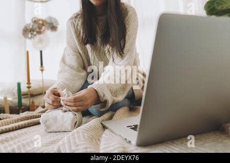 Confezione regalo Furoshiki. Donna che avvolge regalo di natale in tessuto di lino primo piano in festoso boho camera decorata con computer portatile, mani da vicino. Zero Wast Foto Stock