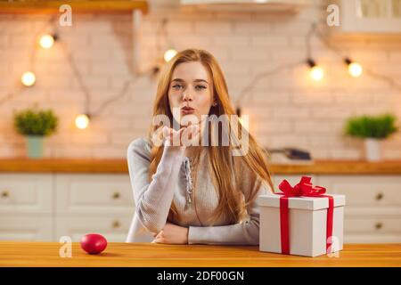 Bella giovane donna seduta al tavolo da cucina con San Valentino presente e soffiare un bacio alla macchina fotografica Foto Stock