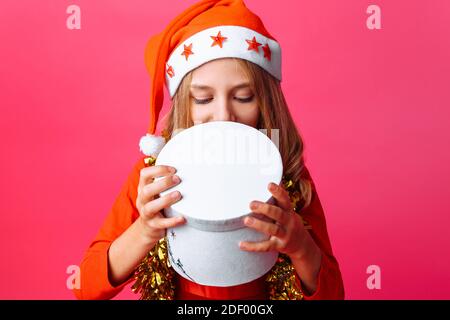 Ragazza teen interessata in cappello di Santa e con tinsel intorno al suo collo peeks con interesse confezione regalo per Natale, su sfondo rosso Foto Stock