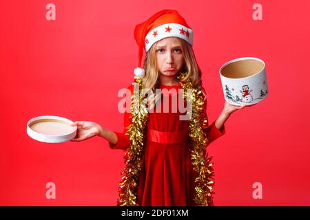 Ragazza teen frustrato in cappello di Santa e con Tinsel sul suo collo, con delusione tenendo casella vuota su sfondo rosso Foto Stock