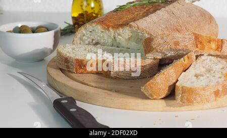 la mano femminile taglia il pane sul tavolo con rosmarino, olive e olio d'oliva. Pane italiano appena sfornato, affettato con un coltello di pane seghettato su un c di legno Foto Stock