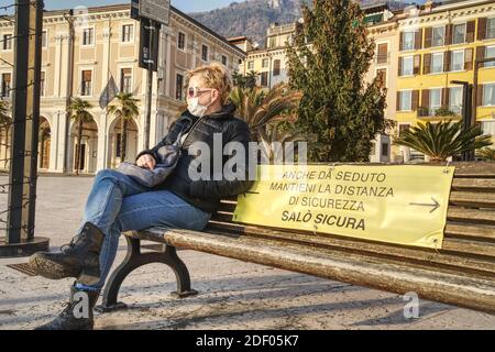 Una donna caucasica di mezza età seduta accanto al segno che dice mentre si siede, mantenere la distanza di sicurezza, salo sicuro o una panca che indossa una protezione del viso m Foto Stock