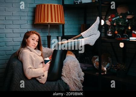 Bella donna con tazza di caffè seduta sulla sedia. Atmosfera di Natale e Capodanno. Tazza di cioccolata calda con marshmallows. Bella ragazza in maglione. Parete grigia in mattoni su sfondo. Vacanza invernale. Foto Stock