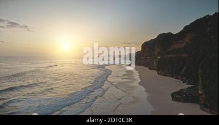 Alba al sole sulle onde dell'oceano con scogliera spiaggia di sabbia di Bawana vista aerea. Paradiso stagcape di natura tropicale con costa rocciosa. Incredibile paesaggio soleggiato di Sumba Island, Indonesia drone shot Foto Stock