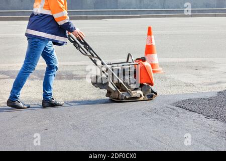 Un operatore stradale compatta l'asfalto sulla carreggiata con un compattatore a piastre vibranti a benzina. Foto Stock