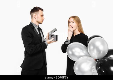 un uomo in costume fa una proposta per il matrimonio con una ragazza che è scioccata, un uomo tiene una scatola con un regalo nelle sue mani davanti al suo volto, su un bianco Foto Stock
