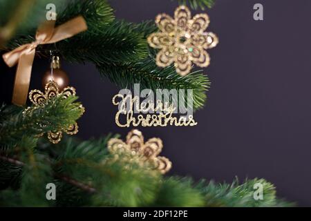 Ornamento Golden albero di Natale con il testo 'buon Natale' appeso ramo di abete con baubles a forma di fiocco di neve sulla schiena scura sfondo Foto Stock