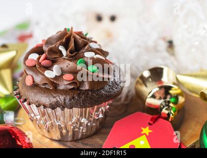 Un cupcake di Natale di cioccolato si siede su un tavolo circondato da etichetta regalo, archi, ornamenti e una Santa. Foto Stock