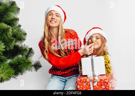 Mamma e figlia indossano cappelli Babbo Natale, nei maglioni di Capodanno, festeggiano il nuovo anno giocando con le palle di Natale. Su sfondo bianco. Foto Stock