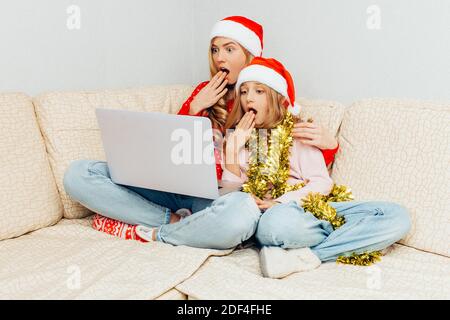 La mamma scioccata e sua figlia, vestita con i cappelli di Babbo Natale, usando un computer portatile mentre si siede sul divano, mostrano emozioni di paura. Foto Stock