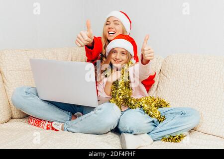 Giovane madre e sua figlia vestite con cappelli di Babbo Natale usando un laptop e sorridendo, seduto sul divano, mostrando una classe di gesti. Foto Stock