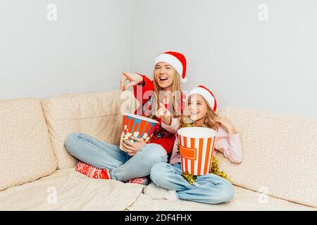 Una giovane madre e sua figlia, vestita di cappelli di Babbo Natale, guardano film e mangiano popcorn mentre si siedono sul divano a casa. Cristo Foto Stock