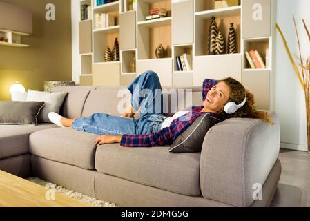 Donna felice in camicia controllata e jeans rilassarsi sul divano in salotto e ascoltando la musica in bianco sopra l'orecchio cuffie Foto Stock
