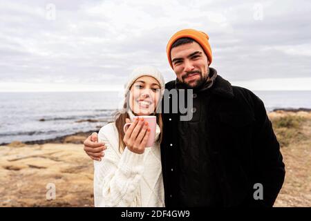 Bel ragazzo abbracciando la sua bella ragazza caucasica che beve un tazza di tè all'aperto in una località balneare in autunno-inverno Colore umore - coupl felice Foto Stock