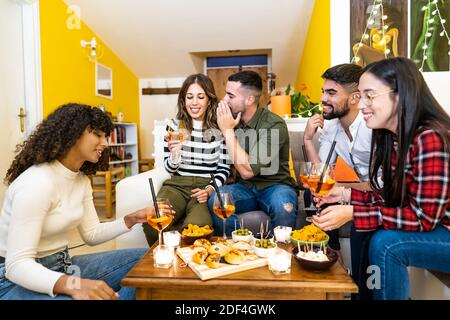 Gruppo di amici di razza mista che si divertono a parlare l'uno con l'altro a casa nel soggiorno seduto sul divano per una festa che beve spritz bevanda lunga coc Foto Stock