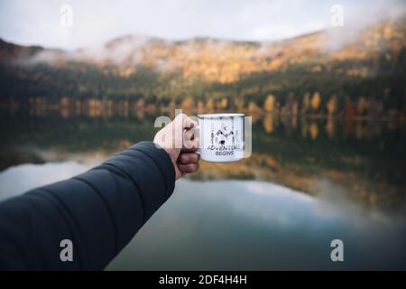 Giovane viaggiatore maschio che tiene una tazza con caffè nella foresta selvaggia, stagione autunnale. Avventura, stile di vita. Foto Stock