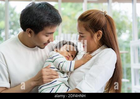 Il padre e la madre asiatici stanno abbracciando 9 mesi il figlio del bambino sul divano. Sorridono e si toccano al bambino con amore nel salotto di casa Foto Stock