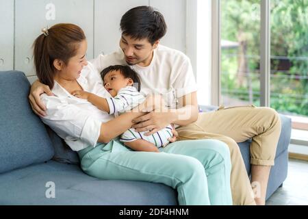 Il padre e la madre asiatici stanno abbracciando 9 mesi il figlio del bambino sul divano. Sorridono e si toccano al bambino con amore nel salotto di casa Foto Stock