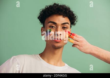 Ritratto di giovane uomo con volto dipinto con bandiera arcobaleno di orgoglio gay. Pittura a mano lgbt colore arcobaleno sul viso di un uomo. Foto Stock