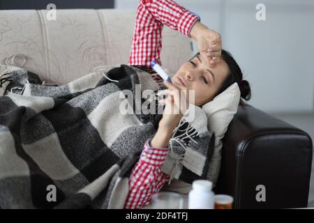 Donna con freddo sdraiato sul divano sotto le coperture e guardando sul termometro Foto Stock