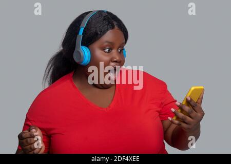 Giovane donna dalla pelle scura con cuffie che tengono uno smartphone e lo sguardo sorpreso Foto Stock