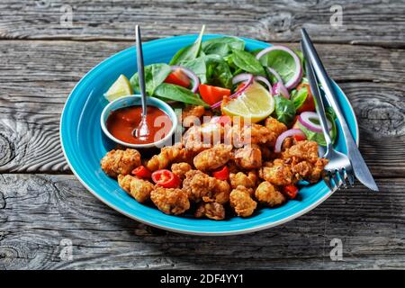 Spuntino americano con pollo al popcorn croccante servito su un piatto blu con ketchup e insalata di spinaci, cipolla rossa, pomodori ciliegini e spicchi di limone su un Foto Stock