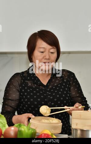 Donna asiatica matura che tiene un gnocco mentre si controlla un tradizionale vapore di bambù con verdure fuori fuoco. Cucina cinese tradizionale concetto. Foto Stock