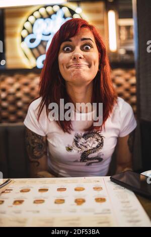 Un ritratto verticale closeup di una donna hipster positiva tatuata con una divertente espressione facciale che si rilassa in un caffè Foto Stock