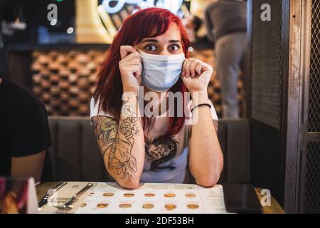 Un ritratto closeup di una donna hipster positiva tatuata dentro una maschera facciale protettiva che si raffredda in un cafe' Foto Stock