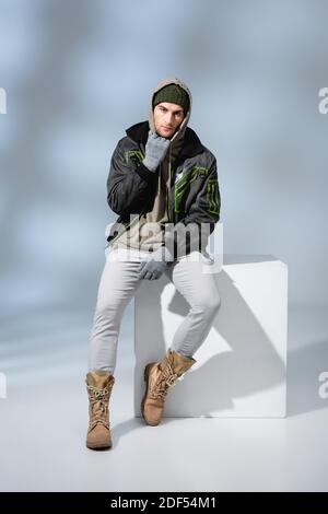 tutta la lunghezza di uomo trendy in cappello, guanti e parka in posa mentre si siede su cubo bianco su grigio Foto Stock