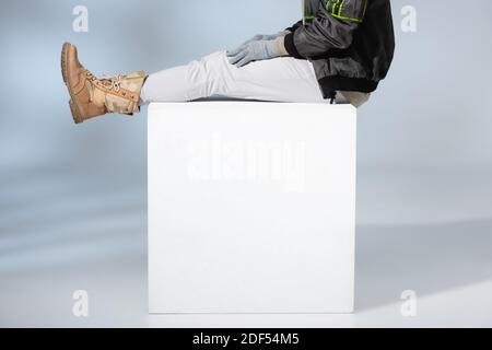 vista ritagliata dell'uomo alla moda in guanti e anorak seduta su cubo bianco su grigio Foto Stock