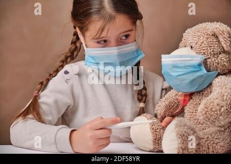 Dottore ragazza in maschera giocando e misurando la temperatura di a. orso giocattolo Foto Stock