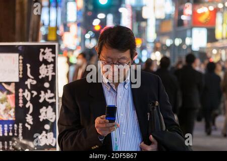 Un salarista o un lavoratore giapponese di sesso maschile controlla il suo telefono cellulare per strada nella zona notte di Shimbashi, Tokyo, Giappone. Foto Stock