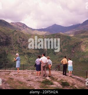 Anni '1960, storico, primaverile e visitatori in alto su un punto panoramico sul Monte Snowdon, Galles, guardando la catena montuosa nel parco nazionale. In quest'epoca, le persone andavano in tali posti con i normali vestiti di tutti i giorni, non in pile sintetiche e stivali da montagna! Foto Stock