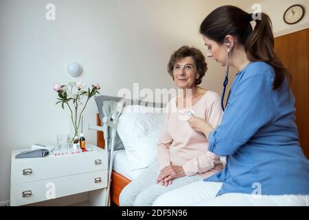 Medico femminile che ascolta i polmoni di paziente anziano con stetoscopio nella stanza dell'ospedale. Home infermiere ascolto per la respirazione di donne anziane. Foto Stock