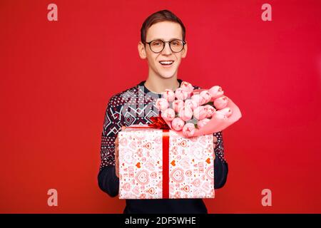 uomo in occhiali, uomo con un dono e un mazzo di fiori in mano, su sfondo rosso Foto Stock