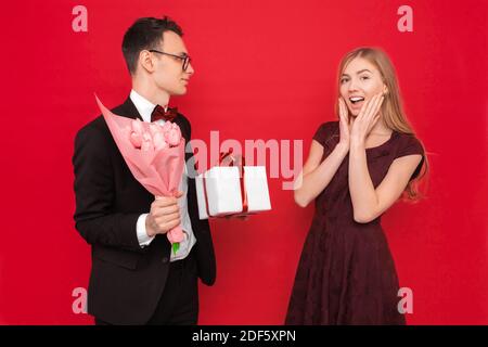 Una coppia amorevole, un uomo dà a una donna scioccata un bouquet di tulipani e una scatola con un regalo su uno sfondo rosso. Concetto di San Valentino Foto Stock