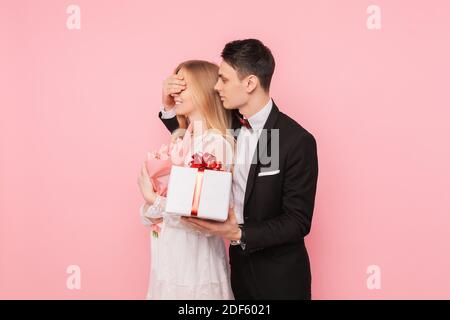 Bell'uomo in un vestito che fa una sorpresa ad una donna, chiudendo gli occhi con le mani su uno sfondo rosa Foto Stock