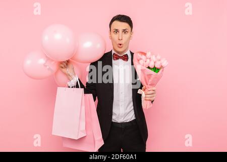 uomo elegante, in costume, con un mazzo di fiori, borse e palloncini, su sfondo rosa Foto Stock