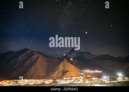 Stepantsminda, Georgia. Cielo stellato di notte con le stelle incandescenti sopra il picco del Monte Kazbek coperto di neve. Villaggio di Kazbegi nella luce notturna Foto Stock
