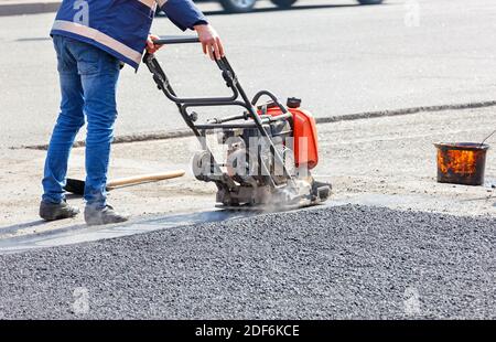 Un addetto alla manutenzione stradale compatta l'asfalto su un tratto stradale recintato della carreggiata con un compattatore a piastre vibranti a benzina. Foto Stock