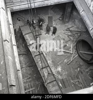 Anni '50, storico, India, Bombay, vista aerea guardando giù la stiva di carico di una nave container e lavoratori indiani che guardano come un grande blocco di metallo pesante è sollevato. Il trasporto di metallo è di due tonnellate in peso e non un elmetto in vista. Alcuni lavoratori portuali sono anche in barefeet o indossare sandali leggeri. Foto Stock