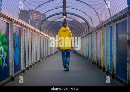 Donna che attraversa un ponte indossando un cappotto giallo, vista posteriore. Foto Stock